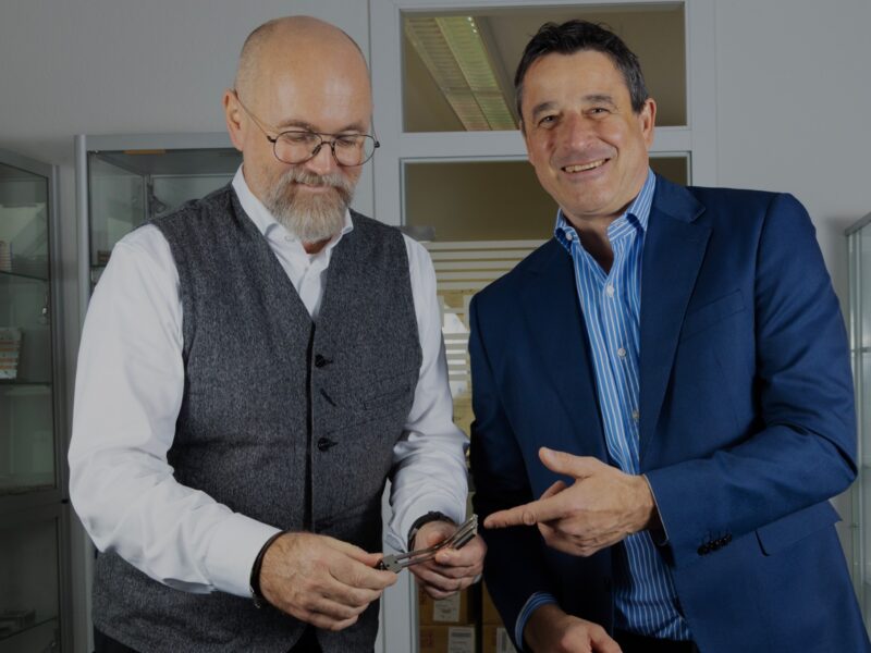 Zwei Männer stehen in einem Büro, der eine hält ein Objekt, der andere zeigt darauf, im Hintergrund sind Regale