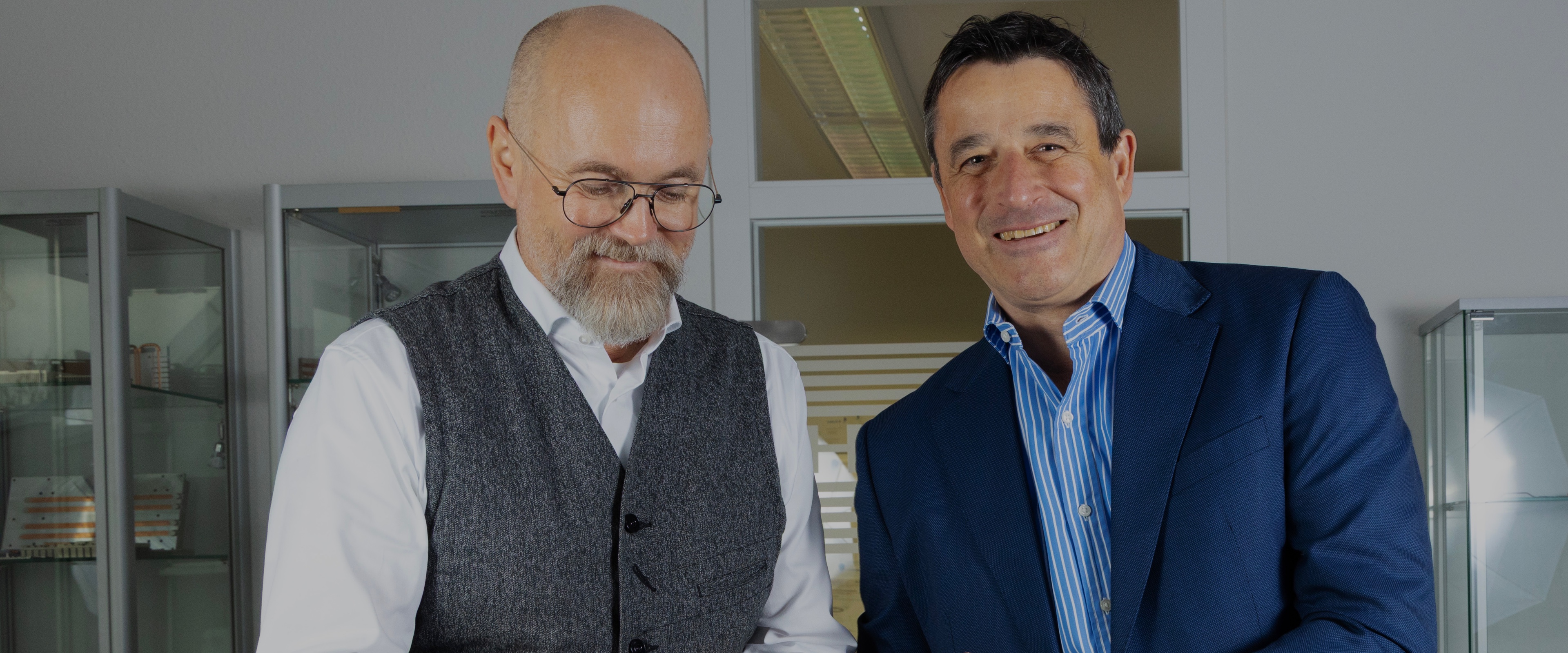 Zwei Männer lächeln in die Kamera, Büro im Hintergrund, Beleuchtung von oben.