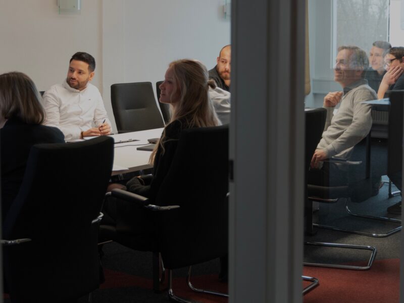 Personen sitzen an einem Konferenztisch in einem Büro und hören einer Präsentation zu.