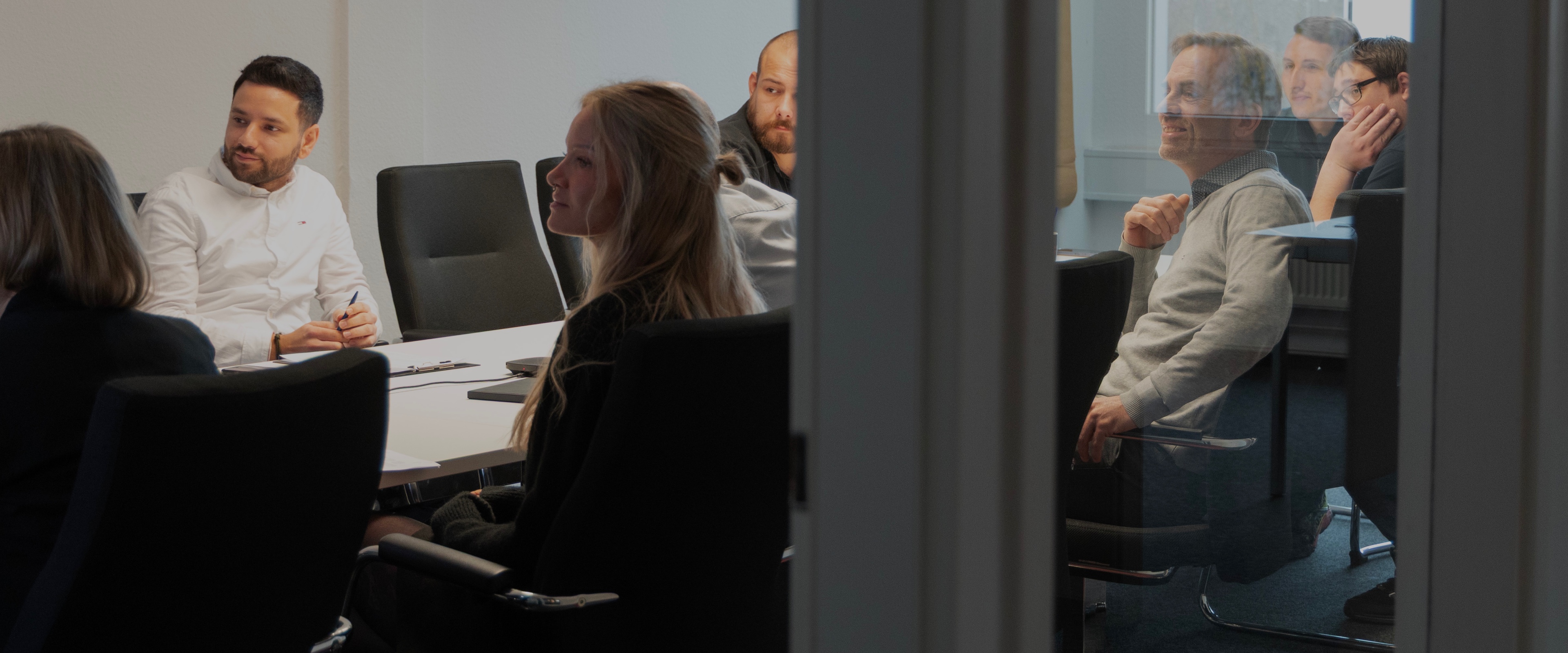 Personen sitzen an einem Besprechungstisch und hören einem Vortrag zu in einem Konferenzraum.
